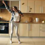 Cheerful young funny woman dancing and singing with ladle while having leisure time in the kitchen at home in the morining